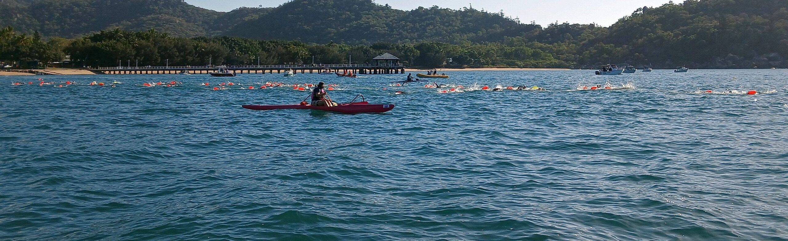 Swimmers mid-crossing with a kayak escort, Picnic Bay jetty in the background