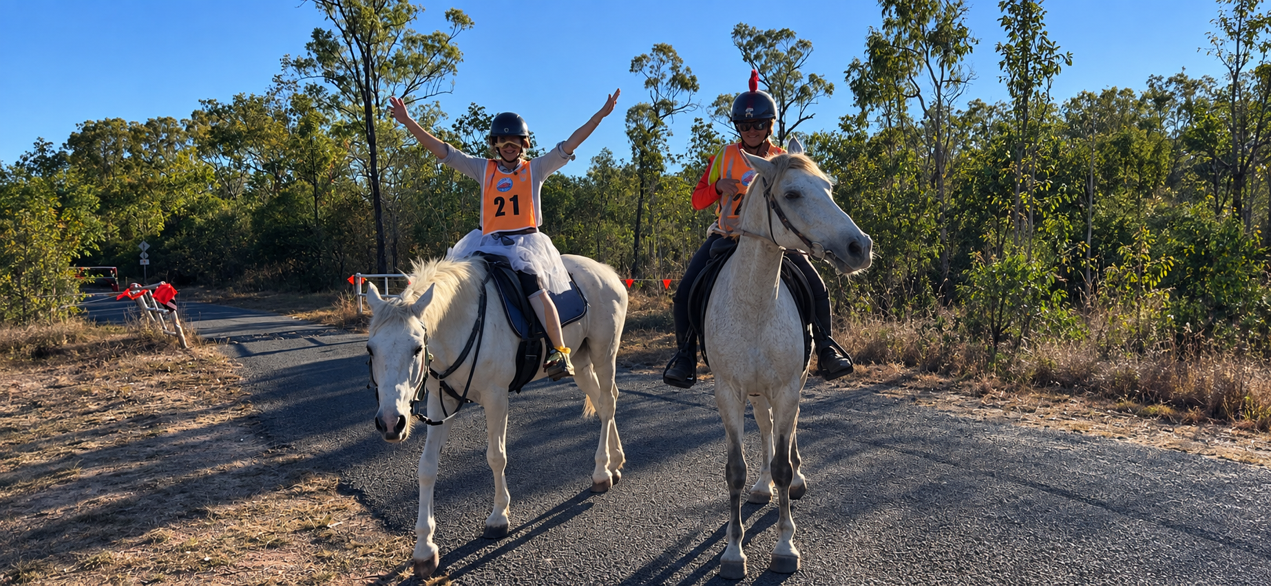 Two riders on white horses on the road, one with arms raised in celebration